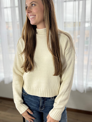 Woman wearing a beige sweater and blue jeans indoors with white curtains in the background