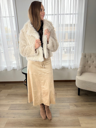 Woman wearing a cream fur coat and beige skirt indoors.