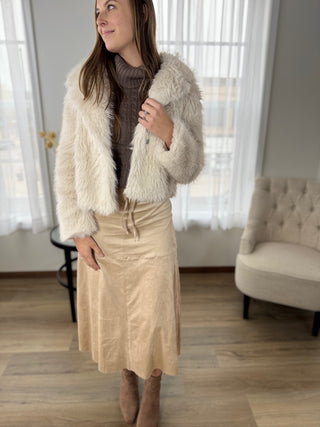 Woman wearing a cream fur coat, brown sweater, and beige skirt in a room with a chair and curtains.