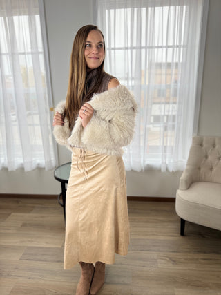 Woman holding a cream-colored fur coat in a room with white curtains and a beige chair.