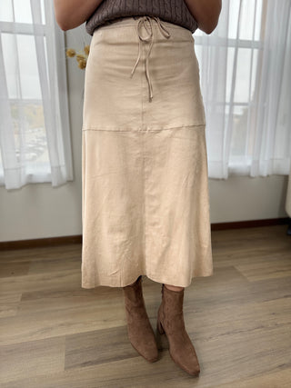 Person wearing a beige skirt and brown boots indoors with white curtains in the background