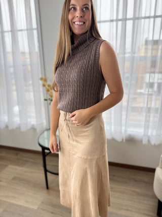 Woman wearing a brown sleeveless knit top and beige skirt indoors.