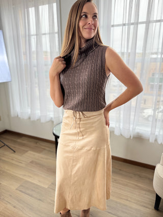 Woman wearing a brown ribbed top and beige skirt indoors with white curtains in the background