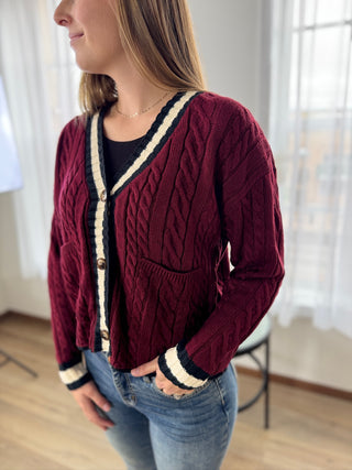 Person wearing a burgundy cable knit cardigan with white and navy trim indoors.