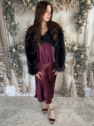 Woman in a burgundy dress with a black fur coat standing in front of a decorated Christmas fireplace.