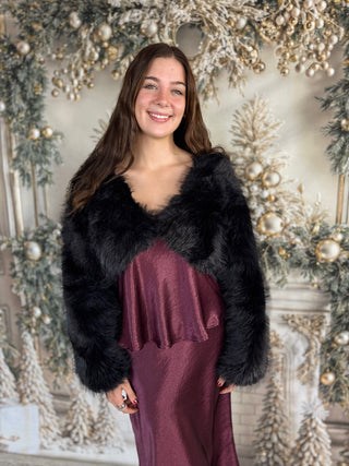 Woman wearing a black fur shrug over a burgundy dress in front of a decorated Christmas background.