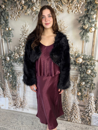 Woman in a burgundy dress with a black fur coat standing in front of a decorated Christmas wall.