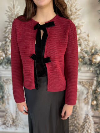 Red knitted jacket with black bow tie worn by a person, against a decorative background.