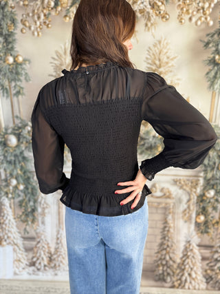 Person wearing a black blouse with sheer sleeves and a textured back against a decorative background.