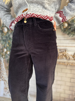Person wearing a gray sweater with red patterned cuffs and dark brown corduroy pants in front of a decorative background.