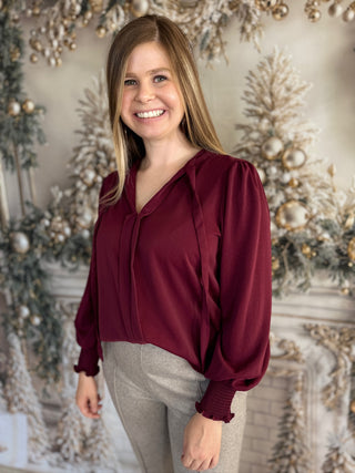 Woman wearing a burgundy blouse and gray pants standing in front of a decorated Christmas background.