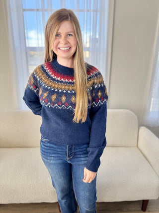 Person wearing a blue patterned sweater standing in a room with a couch and window.