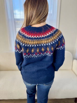 Person wearing a blue sweater with colorful patterns in a room with white curtains.