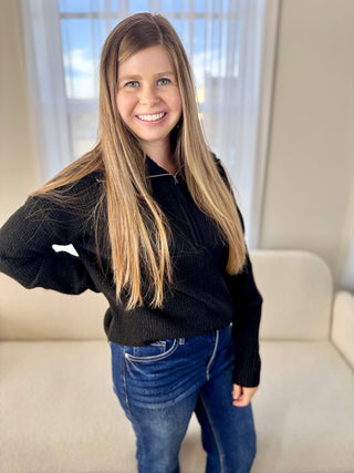 Woman wearing a black sweater and blue jeans standing in a room with a couch and window.