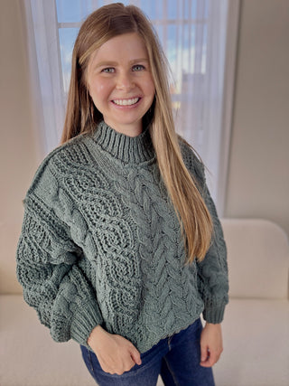 Woman wearing a green cable knit sweater in a room with a window in the background
