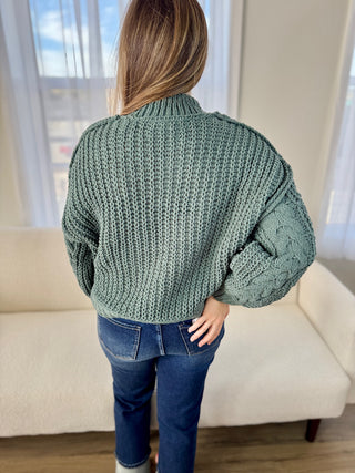 Person wearing a green chunky knit sweater and blue jeans indoors.
