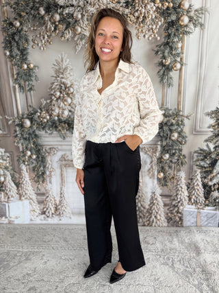 Woman in a white lace blouse and black pants standing in front of a decorated Christmas wall.