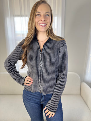 Woman wearing a gray ribbed sweater and blue jeans indoors.