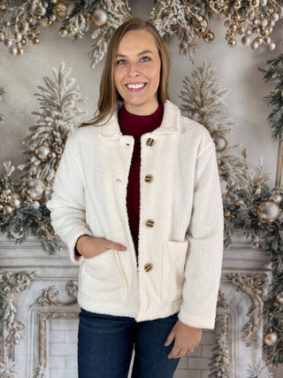 Woman wearing a cream sherpa jacket in front of a decorated fireplace.