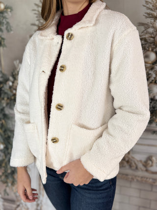 Person wearing a white sherpa jacket with gold buttons against a decorative background.