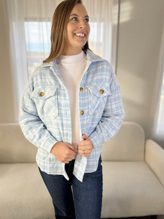 Woman wearing a light blue plaid shirt over a white sweater in a room with a couch and window.