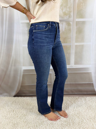 A person wearing dark blue high-rise flare jeans, paired with a cream-colored top, standing barefoot on a white shag rug in front of a window with sheer curtains.