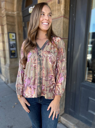 A woman wearing a long-sleeve V-neck blouse with a pink and beige floral pattern, paired with dark blue jeans, standing outdoors in front of a building entrance.