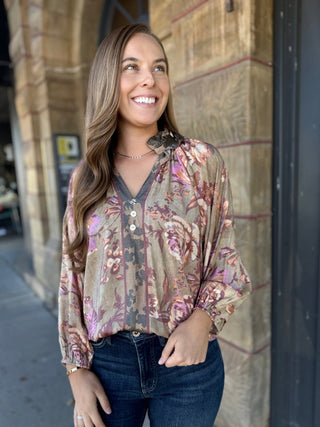 A woman smiling and looking upward while wearing a long-sleeve floral blouse with a V-neck and dark blue jeans, standing outdoors against a stone building.