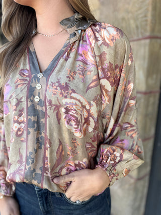 A woman wearing a long-sleeve V-neck blouse with a floral print in muted beige, mauve, and brown tones, paired with dark blue jeans.