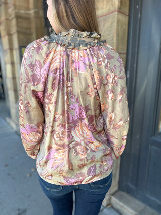 A woman viewed from behind wearing a beige long-sleeve blouse with a pink and purple floral pattern and a ruffled high neckline, paired with dark blue jeans.