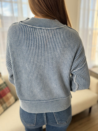 Person wearing a gray ribbed sweater in a room with light-colored curtains.