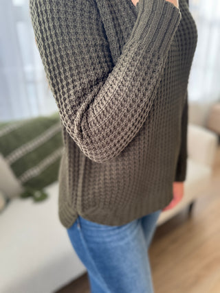 Person wearing a brown knitted sweater and blue jeans in an indoor setting