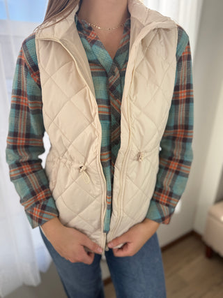 Person wearing a beige quilted vest over a plaid shirt with a white background