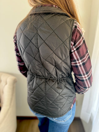 Person wearing a gray quilted vest over a plaid shirt indoors.
