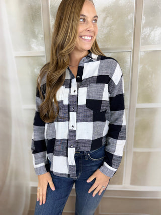 A woman wearing a black, white, and gray buffalo plaid button-down shirt with dark blue jeans, standing in front of a soft-focus window.