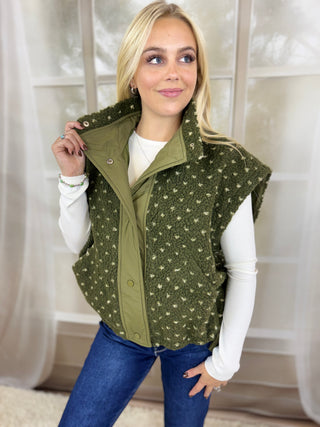 A woman wearing an olive green sleeveless fleece vest with a small white floral pattern, layered over a white long-sleeve shirt and blue jeans, standing in front of a soft-focus indoor background.