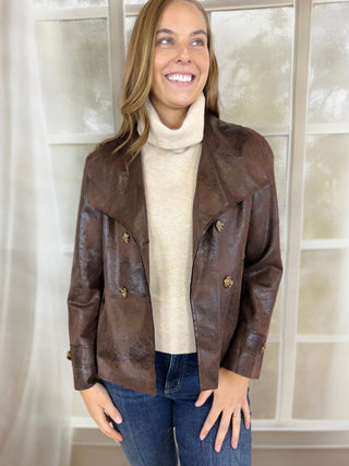 A woman wearing a brown distressed leather jacket over a cream turtleneck sweater and blue jeans, standing in front of a white paneled window.