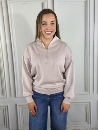 Woman wearing a light pink pullover with a quarter zip and blue jeans against a white paneled wall.