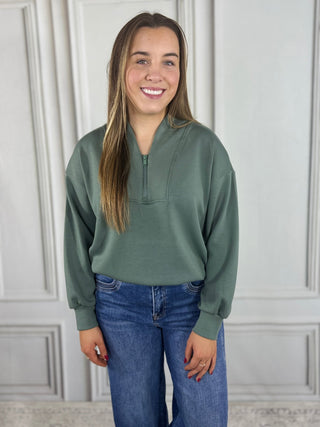 Woman wearing a green sweatshirt and blue jeans standing against a white paneled wall.