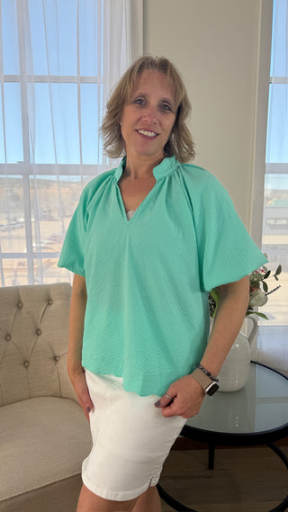 Woman wearing a green blouse and white shorts standing in a room with large windows.