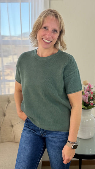 Woman wearing a green sweater and blue jeans standing in a room with a couch and window.