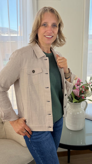 Woman wearing a beige jacket and blue jeans indoors with a table and flowers in the background