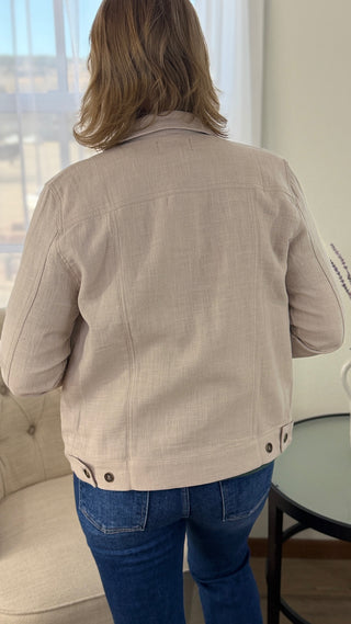 Person wearing a beige jacket and blue jeans in a room with a window and chair.