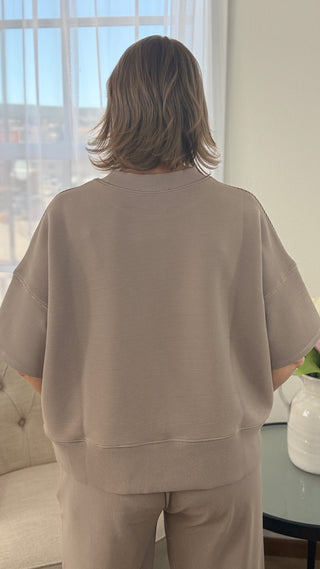 Person wearing a beige outfit standing in a room with a window in the background