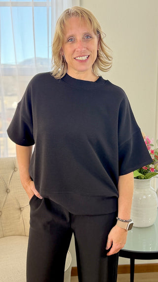 Person wearing a black oversized shirt and pants in a room with a couch and window.