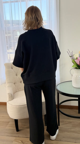 Person wearing a black outfit standing in a room with a chair and table.