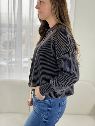 Woman wearing a dark gray ribbed sweater and blue jeans in a bright room.