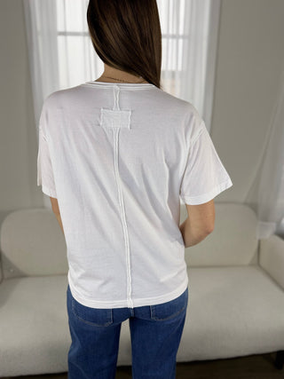 Person wearing a white t-shirt with a small square logo on the back, standing in a room with a couch and curtains.