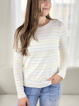 Woman wearing a white sweater with colorful stripes and blue jeans indoors.