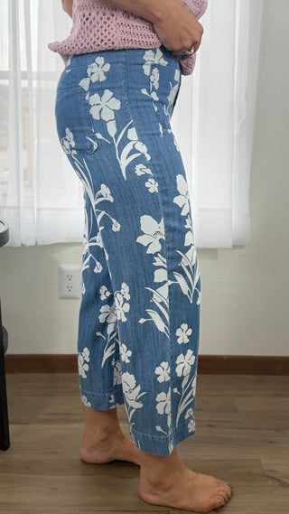 Person wearing blue floral pants standing indoors with white curtains in the background